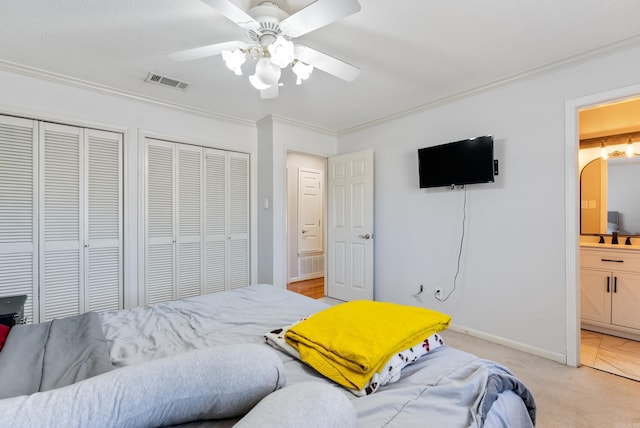 bedroom featuring visible vents, light colored carpet, ornamental molding, ensuite bathroom, and two closets