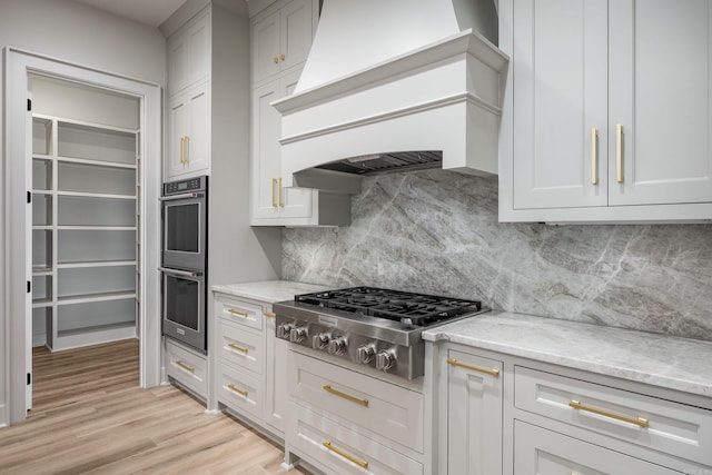 kitchen featuring decorative backsplash, light stone counters, custom exhaust hood, stainless steel appliances, and light wood-style floors