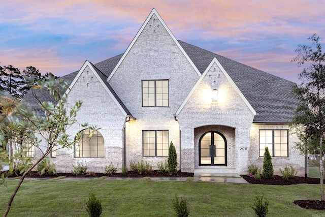 view of front facade with roof with shingles, brick siding, a lawn, and french doors