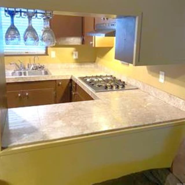 kitchen featuring under cabinet range hood, a sink, brown cabinetry, light countertops, and stainless steel gas cooktop