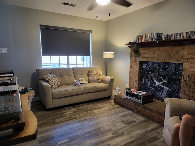 living room with a textured ceiling, wood finished floors, visible vents, a ceiling fan, and a brick fireplace