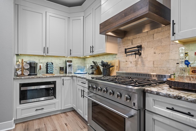 kitchen with appliances with stainless steel finishes, custom exhaust hood, light stone countertops, light wood finished floors, and tasteful backsplash