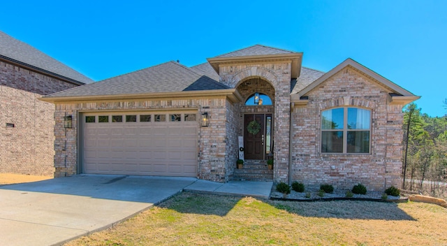 french country inspired facade featuring a shingled roof, concrete driveway, brick siding, and an attached garage