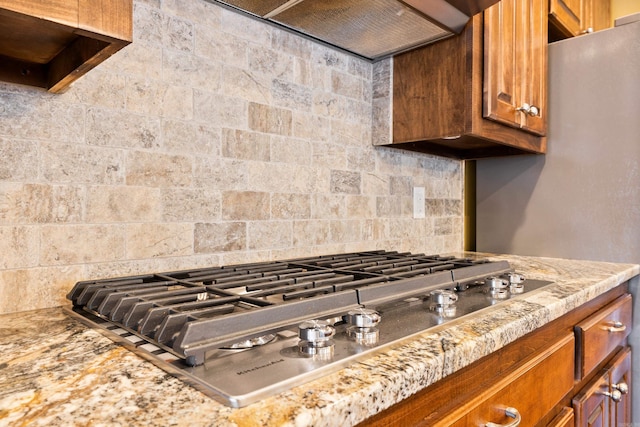 kitchen with ventilation hood, backsplash, light stone countertops, brown cabinetry, and stainless steel gas stovetop