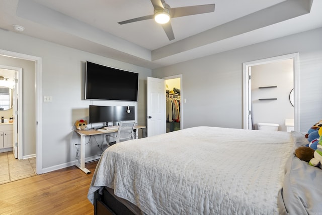 bedroom with baseboards, a walk in closet, a raised ceiling, light wood finished floors, and ensuite bath