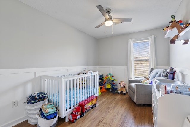 bedroom with a crib, wainscoting, wood finished floors, and a ceiling fan