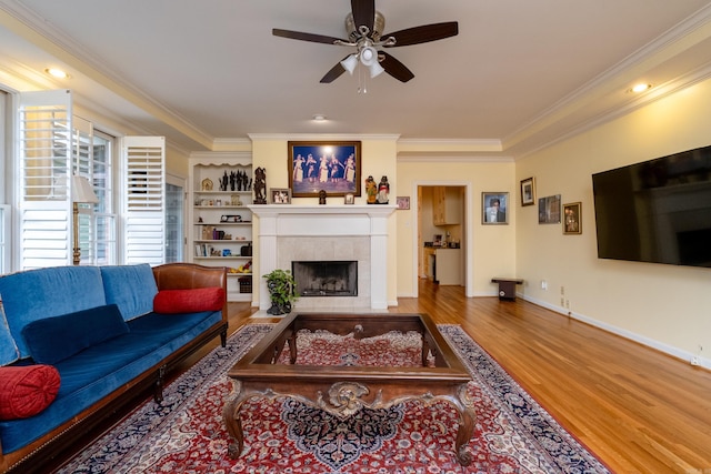 living area featuring ceiling fan, wood finished floors, baseboards, a tiled fireplace, and crown molding