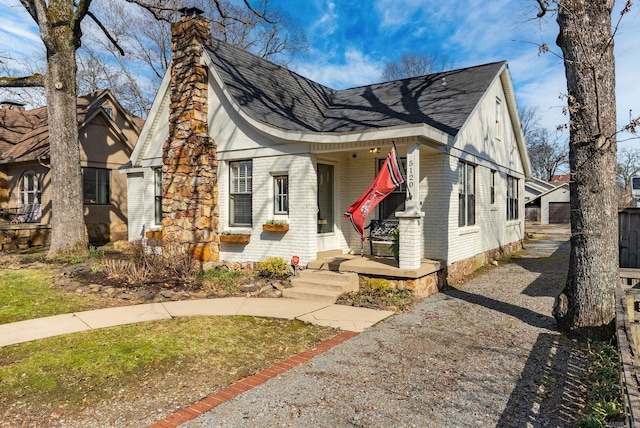 tudor home featuring brick siding, a porch, a chimney, and roof with shingles