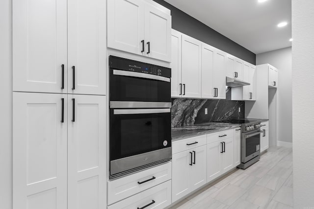 kitchen featuring multiple ovens, under cabinet range hood, dark stone counters, decorative backsplash, and high end stainless steel range oven