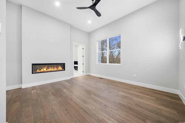 unfurnished living room featuring a glass covered fireplace, ceiling fan, baseboards, and wood finished floors