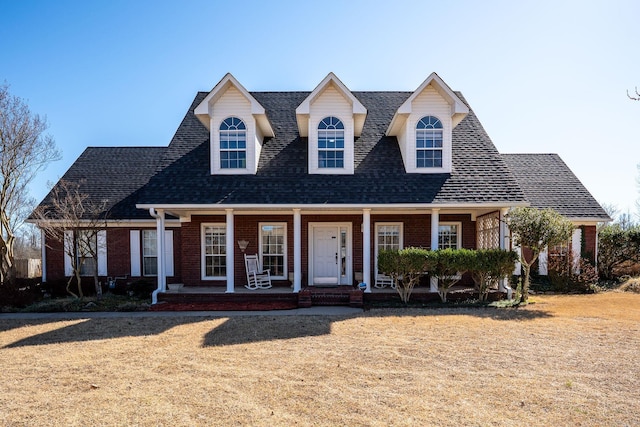 cape cod house featuring brick siding, covered porch, a front lawn, and a shingled roof