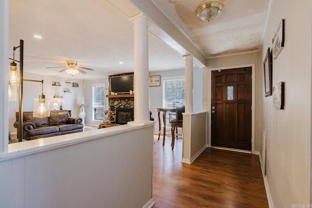 foyer with crown molding, dark wood finished floors, ornate columns, a stone fireplace, and a ceiling fan