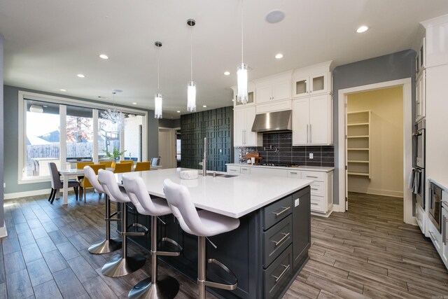 kitchen with ventilation hood, light countertops, decorative backsplash, a large island with sink, and white cabinetry