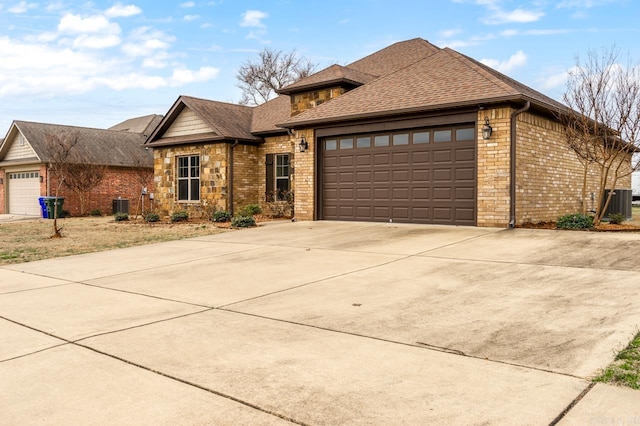 view of front facade with cooling unit, driveway, a shingled roof, stone siding, and a garage