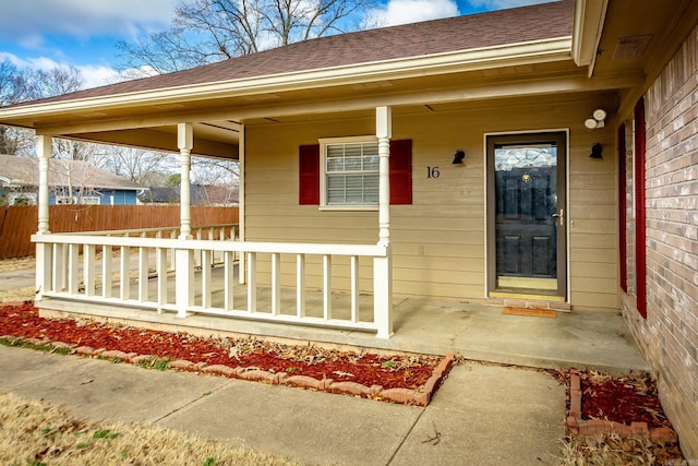 entrance to property featuring brick siding, covered porch, a shingled roof, and fence