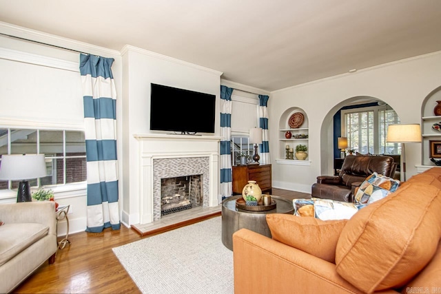 living area featuring a fireplace with raised hearth, built in shelves, crown molding, wood finished floors, and arched walkways