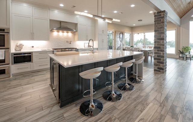 kitchen with decorative backsplash, a sink, wall chimney exhaust hood, light countertops, and decorative columns