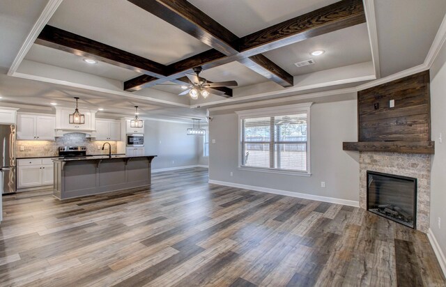 unfurnished living room featuring beam ceiling, a ceiling fan, wood finished floors, a stone fireplace, and baseboards