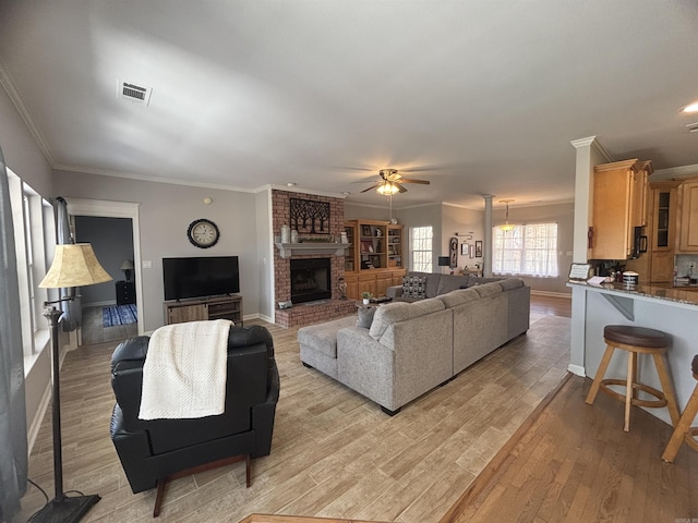 living area with visible vents, light wood finished floors, ceiling fan, crown molding, and a brick fireplace