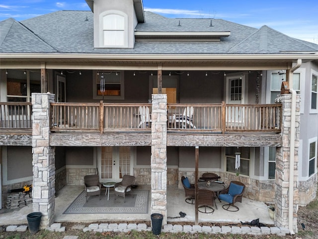 back of house with stone siding, a balcony, roof with shingles, and a patio