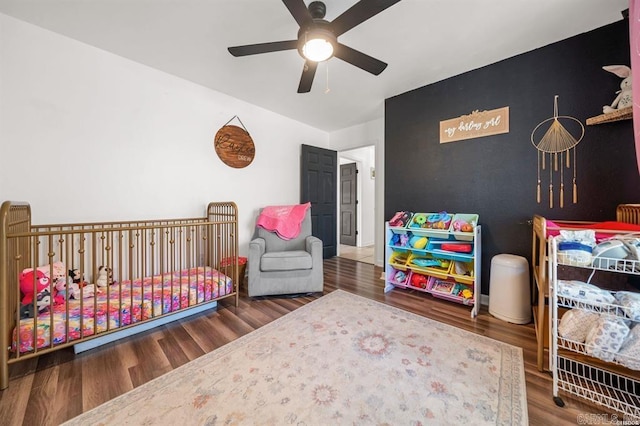 bedroom with a crib, a ceiling fan, wood finished floors, and an accent wall