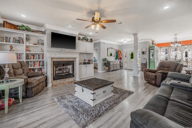 living room featuring ornate columns, a tile fireplace, light wood-style floors, crown molding, and ceiling fan with notable chandelier