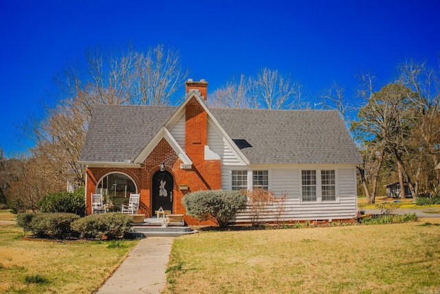 tudor-style house featuring brick siding, a chimney, a front yard, and a shingled roof