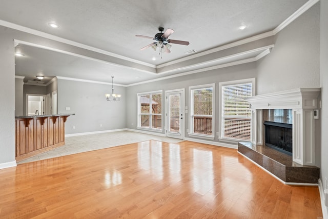 unfurnished living room with ornamental molding, ceiling fan with notable chandelier, a tray ceiling, a high end fireplace, and light wood finished floors