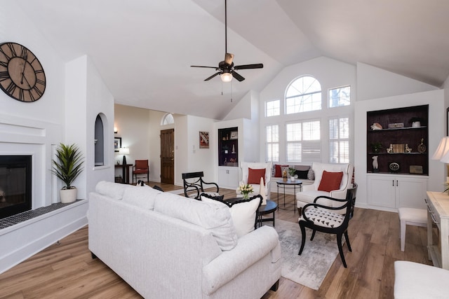 living area with high vaulted ceiling, a ceiling fan, built in features, wood finished floors, and a glass covered fireplace