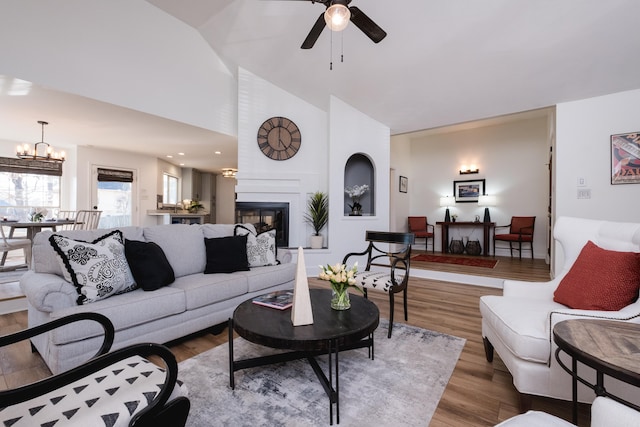 living area with ceiling fan with notable chandelier, high vaulted ceiling, wood finished floors, and a large fireplace