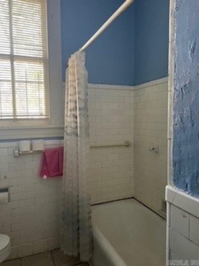 bathroom with a wainscoted wall, toilet, shower / bathtub combination with curtain, tile patterned flooring, and tile walls