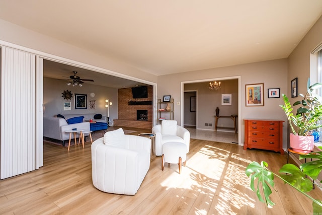 living area with ceiling fan with notable chandelier, a brick fireplace, and light wood finished floors