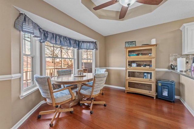 dining room with a ceiling fan, wood finished floors, baseboards, a tray ceiling, and a wood stove