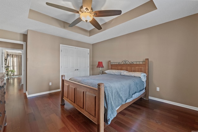 bedroom with baseboards, a closet, a tray ceiling, dark wood-style floors, and a ceiling fan