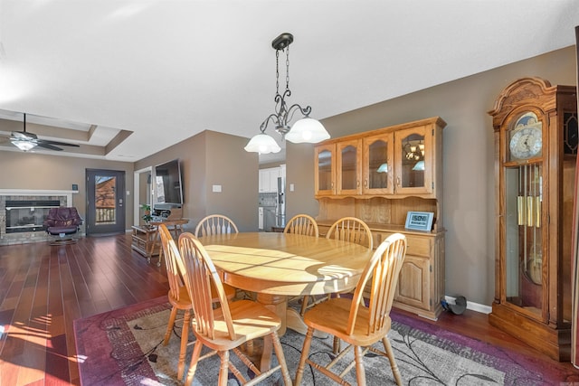 dining space featuring a tray ceiling, a glass covered fireplace, wood finished floors, baseboards, and ceiling fan