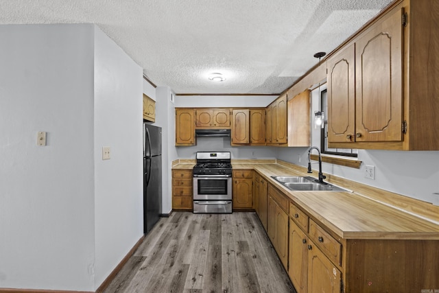 kitchen featuring stainless steel range with gas stovetop, freestanding refrigerator, exhaust hood, brown cabinetry, and a sink