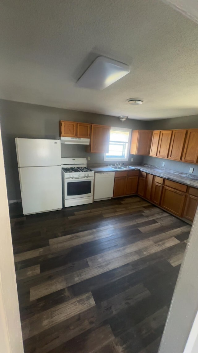 kitchen with under cabinet range hood, a textured ceiling, dark wood-style floors, white appliances, and brown cabinetry