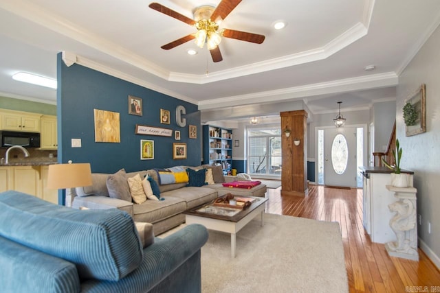 living room with light wood-type flooring, baseboards, crown molding, a raised ceiling, and ceiling fan