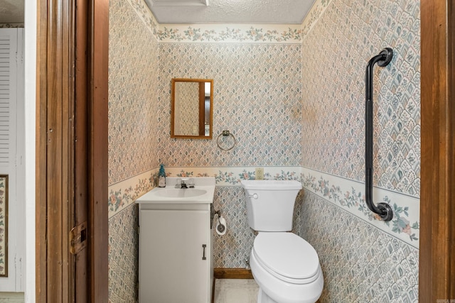 half bath with a textured ceiling, toilet, vanity, and wallpapered walls