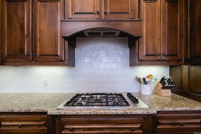 kitchen featuring under cabinet range hood, light stone counters, backsplash, dark brown cabinetry, and stainless steel gas stovetop