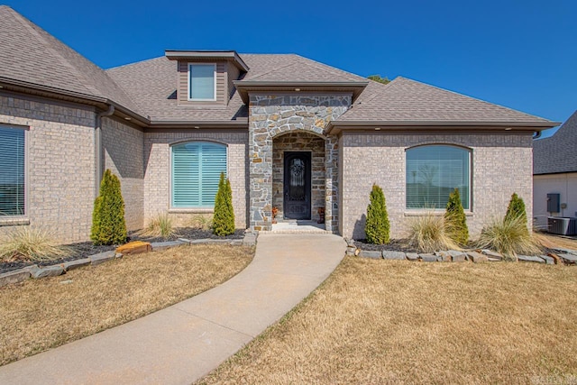 french country home with central air condition unit, brick siding, roof with shingles, and a front lawn