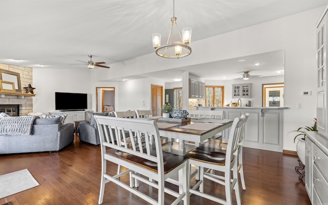 dining space featuring a stone fireplace, recessed lighting, ceiling fan with notable chandelier, and dark wood-style flooring