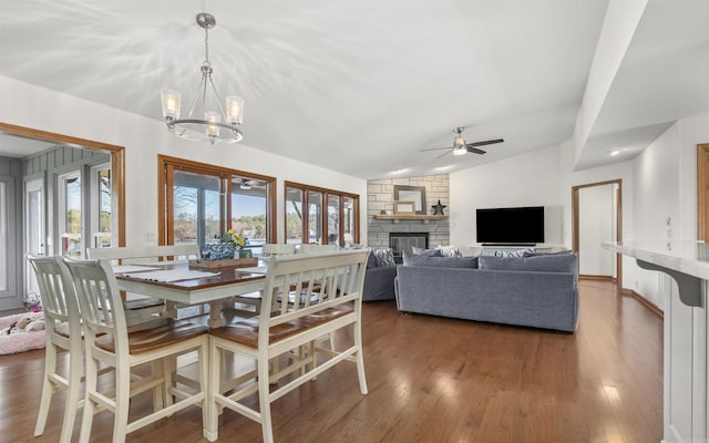 dining space featuring lofted ceiling, a stone fireplace, dark wood finished floors, and ceiling fan with notable chandelier