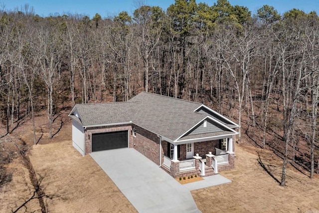 view of front of home featuring brick siding, a porch, a view of trees, driveway, and an attached garage