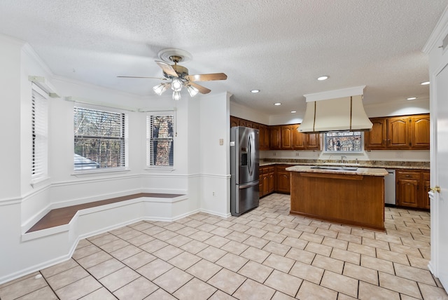 kitchen featuring brown cabinetry, a ceiling fan, a kitchen island, custom exhaust hood, and stainless steel refrigerator with ice dispenser