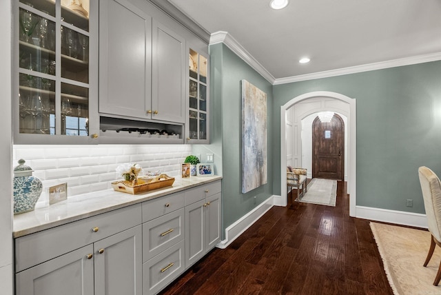 interior space with tasteful backsplash, glass insert cabinets, crown molding, dark wood finished floors, and arched walkways