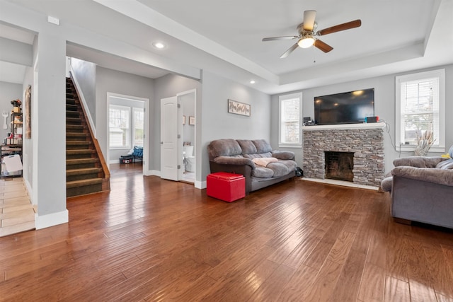 living area featuring stairway, baseboards, ceiling fan, a stone fireplace, and hardwood / wood-style flooring