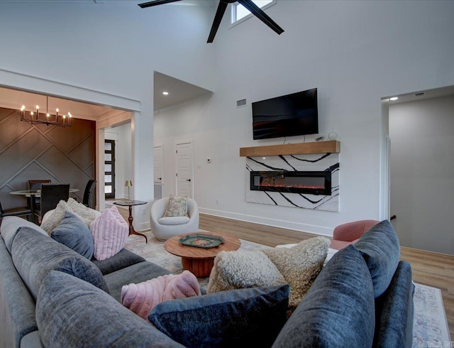 living area featuring wood finished floors, visible vents, a high ceiling, a glass covered fireplace, and ceiling fan with notable chandelier