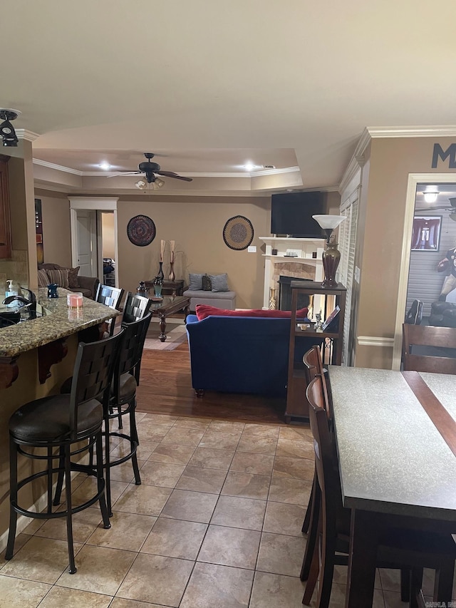 dining area featuring light tile patterned floors, a multi sided fireplace, ceiling fan, crown molding, and a raised ceiling