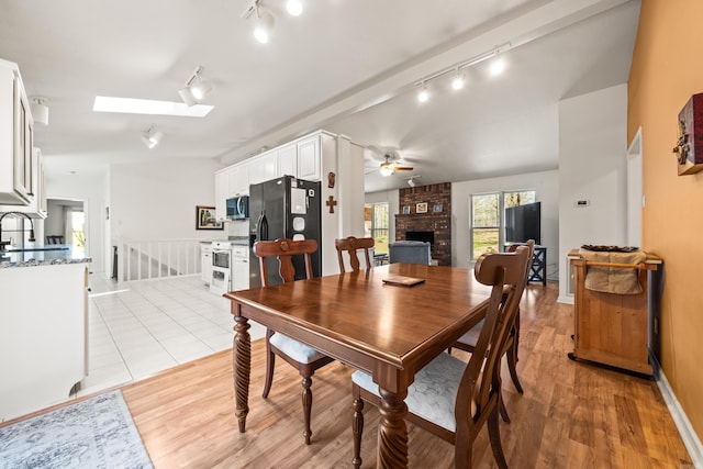 dining room with light wood finished floors, a skylight, ceiling fan, rail lighting, and a brick fireplace